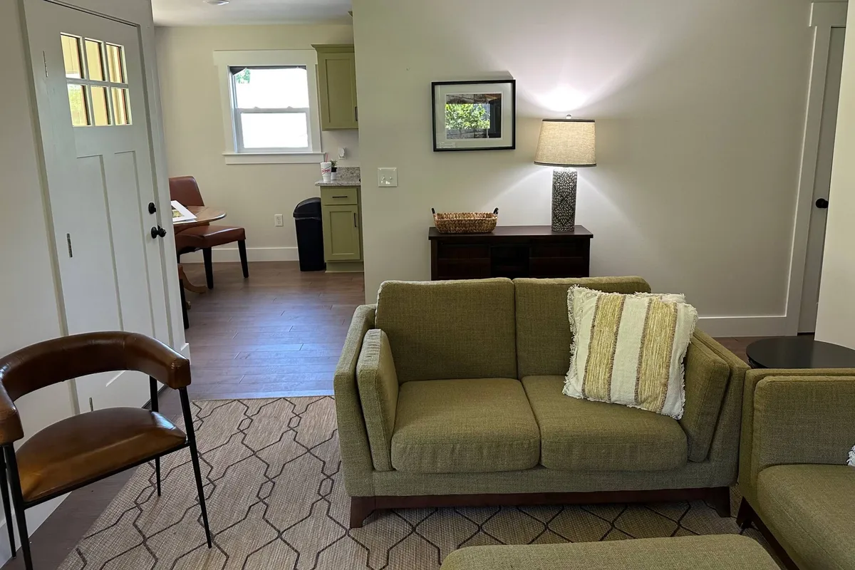 A cozy and stylish living zone anchored by a green sofa with a striped cushion, a table lamp, a wooden cabinet, and a visible dining area beyond. The ensemble creates an inviting atmosphere for relaxing, working, or socializing.