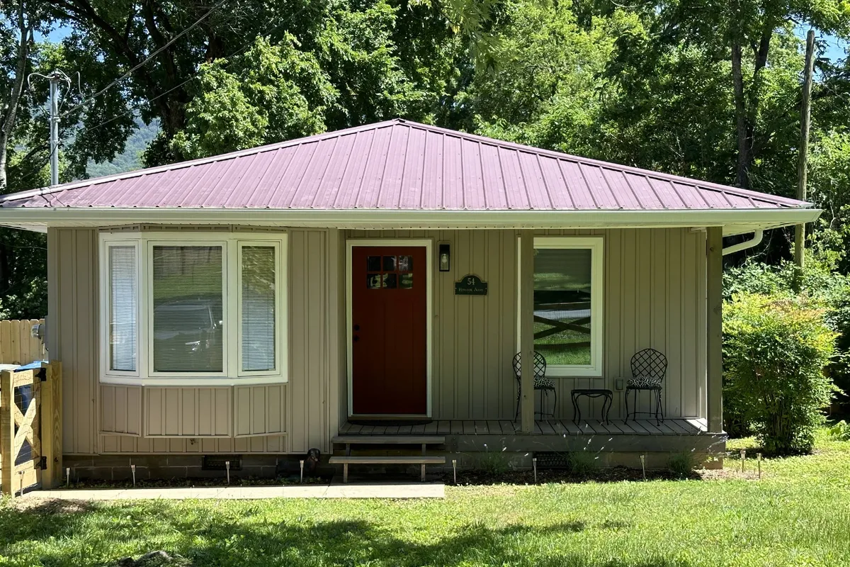 The image features a welcoming front porch with a bright red door, two wrought iron chairs, large windows, and a metal roof. The setup suggests a cozy space for sipping coffee while enjoying the neighborhood activity and garden view.