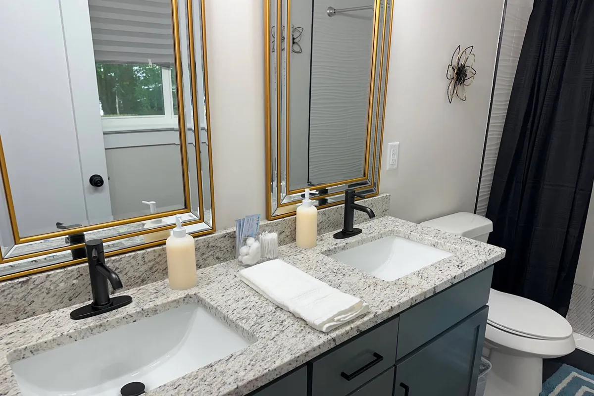 A modern bathroom with double sinks, gold-framed mirrors, and a granite countertop. The space is bright, clean, and designed for convenience, offering an inviting area for morning or evening routines.