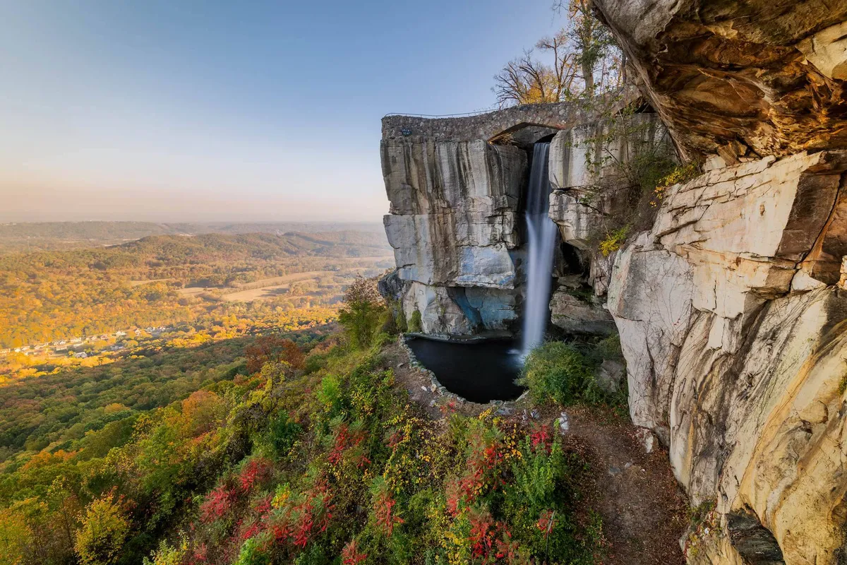 A stunning waterfall cascades over natural rock formations, surrounded by vibrant autumn foliage and offering a sweeping view of the valley below. The scene captures the tranquility and rugged beauty of the outdoors, with hints of nearby hiking trails and spots ideal for photography and picnicking.
