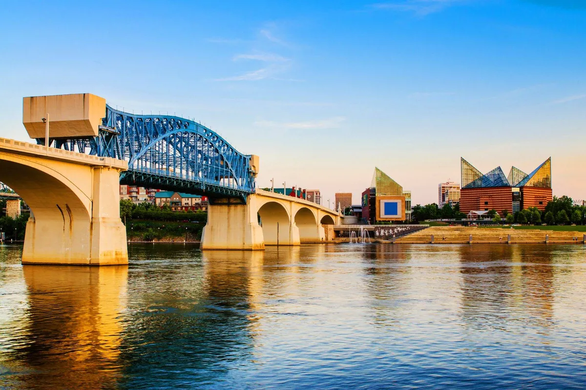 A scenic vista of downtown Chattanooga featuring a striking blue bridge crossing the river, with modern buildings lining the waterfront and their reflections shimmering on the water. The scene is bathed in soft natural light, inviting exploration of the city's vibrant atmosphere and outdoor spaces.