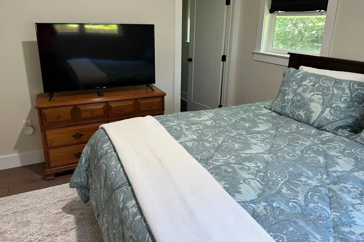 This image shows a serene bedroom featuring a bed with patterned bedding, a wooden dresser with a television, and soft natural lighting. The arrangement suggests a restful environment for unwinding after a full day.