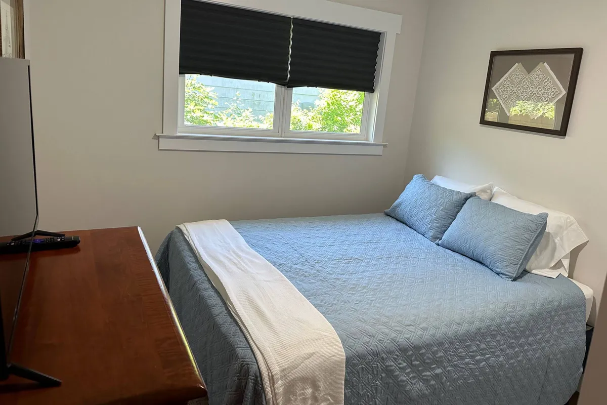 A serene bedroom scene featuring a bed with inviting blue bedding, a sturdy wooden dresser, and a flat-screen television. Soft morning light filters in through the window, casting a tranquil glow across the room, suggesting comfort and privacy ideal for evening relaxation or a restful sleep.