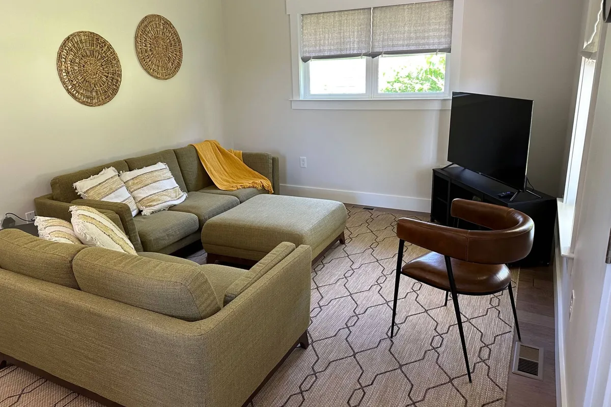 The image depicts a cozy living room with a comfortable sofa, a television, decorative wall hangings, and a chair arranged for relaxation and socializing. Natural light fills the space, creating a warm and inviting atmosphere suited for gatherings with family or friends.