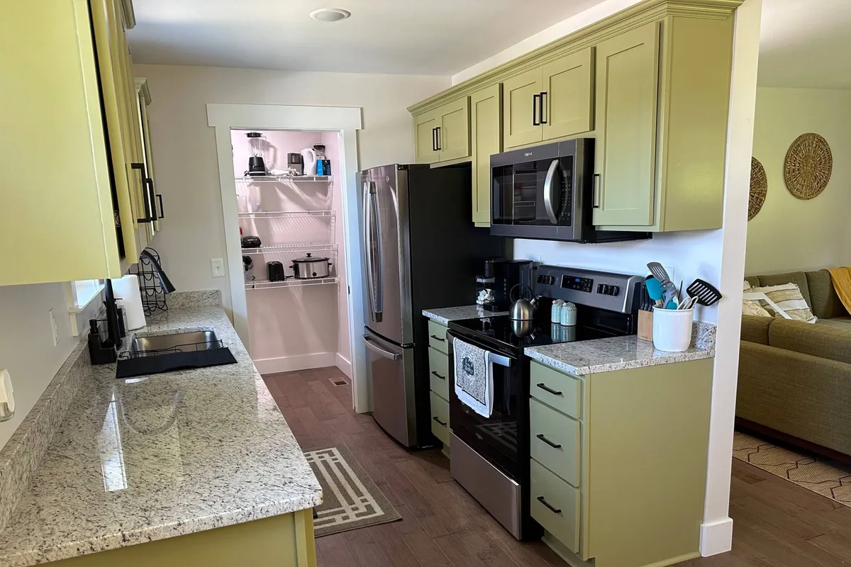 A welcoming kitchen space featuring gleaming granite countertops, stainless steel appliances, and stylish green cabinetry. An open pantry can be seen just off to the side, and the counters are spacious, allowing for meal prep or baking activities. The room exudes a cozy yet highly functional vibe, perfect for family cooking or entertaining guests.