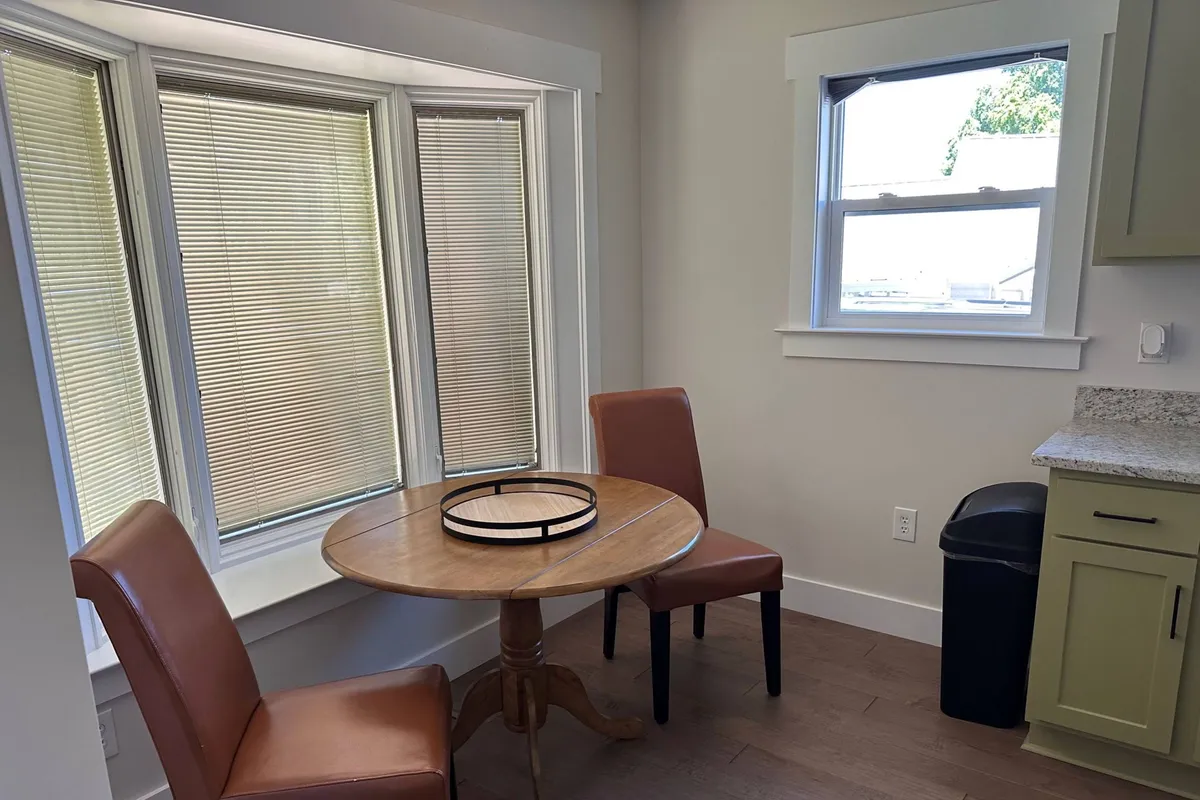 A sun-drenched dining nook with a round wooden table and two upholstered chairs placed near large windows with blinds. The abundant natural light streams in, creating an inviting spot ideal for meals, reading, or working by the window.
