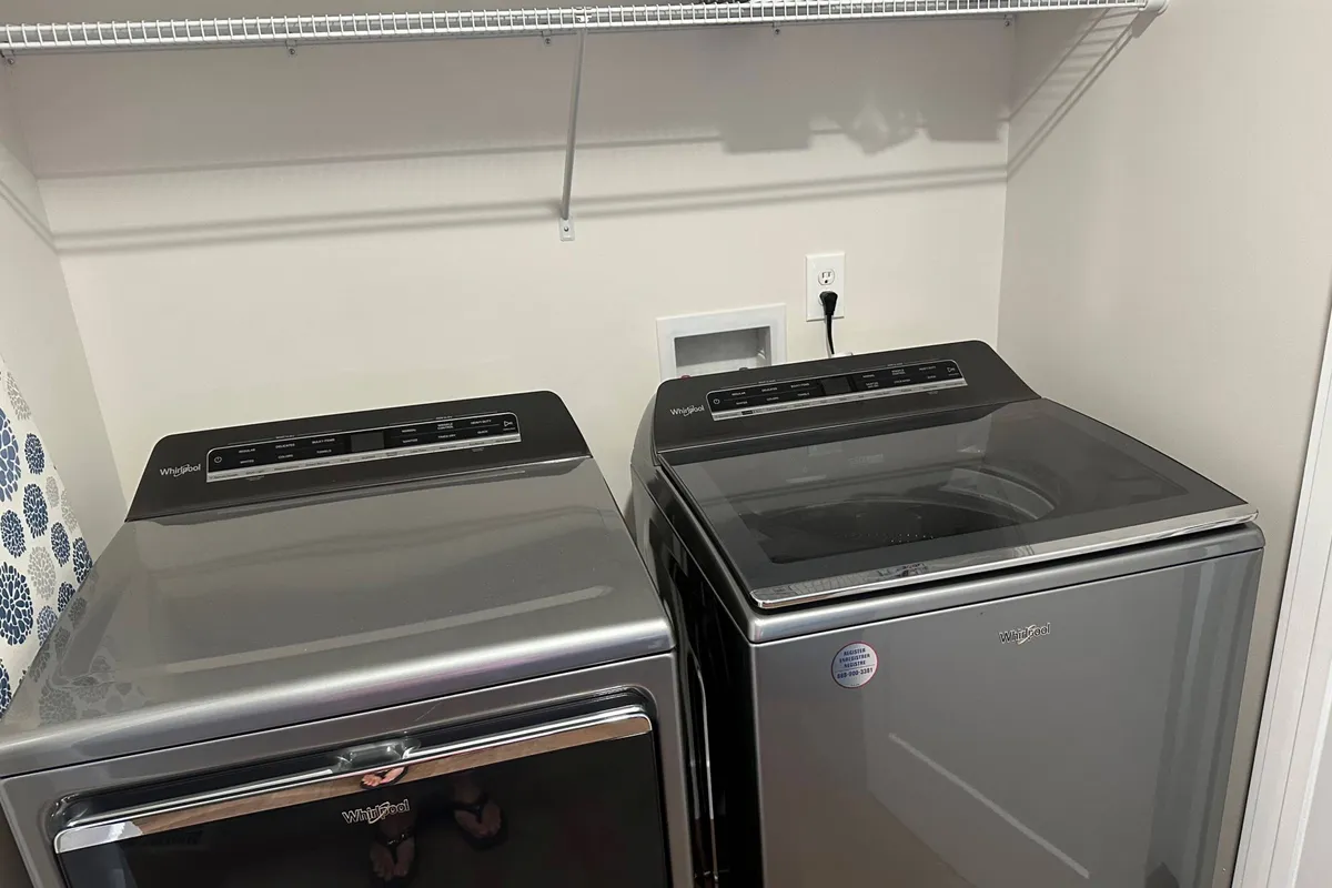 The image shows an organized interior laundry space with a washer, dryer, and shelf. The mood is practical, and it highlights easy-maintenance interiors and satisfaction in everyday routines.